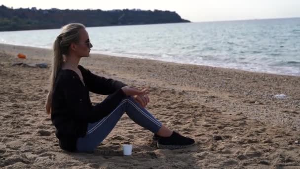 En vacker blondin med långt hår sitter på stranden. Hon är klädd i jeans och en mörk tröja, kastar småsten i havet. Begreppet fritid, resor. Högkvalitativ 4k-film. — Stockvideo