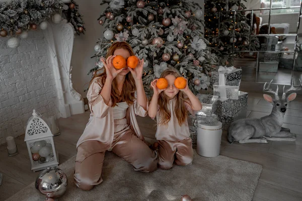 Η μαμά και η κόρη κρατούν μανταρίνια στο ύψος των ματιών αντί για γυαλιά. Κάθονται κάτω από ένα στολισμένο χριστουγεννιάτικο δέντρο με τις πιτζάμες τους. Η έννοια των Χριστουγέννων, οικογενειακές διακοπές — Φωτογραφία Αρχείου