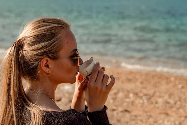 Una hermosa rubia con el pelo largo reunida en una cola de caballo se sienta en la playa bebiendo café y disfrutando de las vistas. — Foto de Stock
