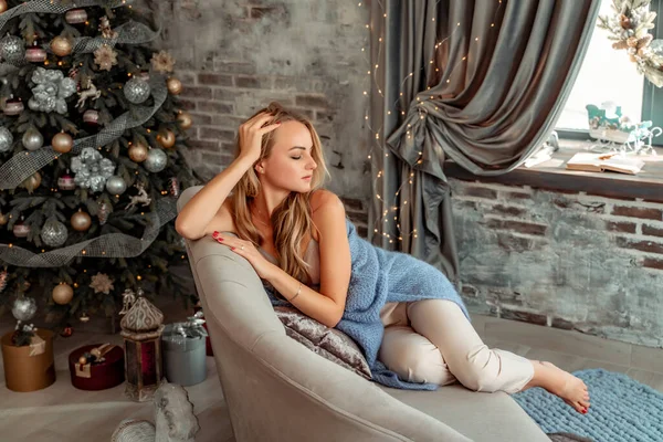 Μια όμορφη ξανθιά κάθεται σε έναν καναπέ σε ένα δωμάτιο με χριστουγεννιάτικες διακοσμήσεις. Χειμερινή διάθεση, άνετο σπίτι, με φόντο ένα διακοσμημένο χριστουγεννιάτικο δέντρο. — Φωτογραφία Αρχείου