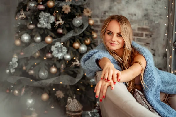 Μια όμορφη ξανθιά κάθεται σε έναν καναπέ σε ένα δωμάτιο με χριστουγεννιάτικες διακοσμήσεις. Χειμερινή διάθεση, άνετο σπίτι, με φόντο ένα διακοσμημένο χριστουγεννιάτικο δέντρο. — Φωτογραφία Αρχείου