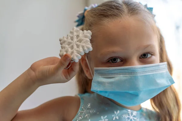 Una chica con una máscara médica sostiene un copo de nieve, quiere decorar el árbol de Navidad. Las vacaciones conceptuales en la pandemia de 2021. — Foto de Stock