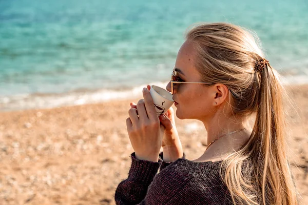 Una hermosa rubia con el pelo largo reunida en una cola de caballo se sienta en la playa bebiendo café y disfrutando de las vistas. — Foto de Stock