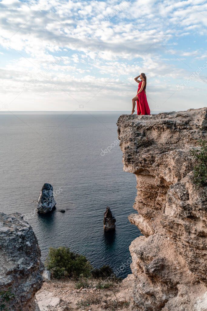 A girl with loose hair in a red dress stands on a rock rock above the sea. In the background, the sea and the rocks. The concept of travel