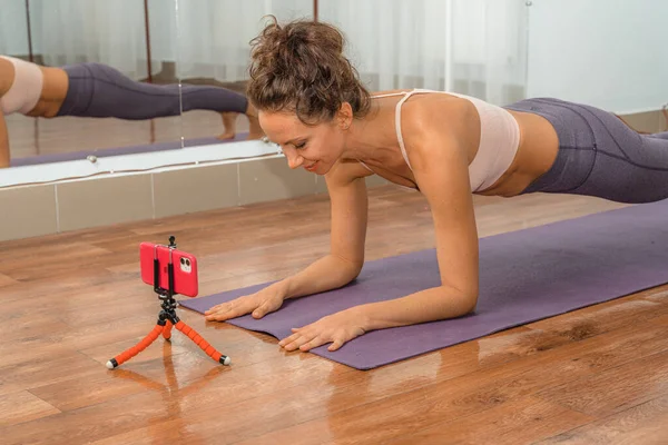 A young beautiful woman practices yoga in a yoga studio and films herself on her phone, watching a lesson. The phone is on a tripod. Harmony, balance, meditation, relaxation, the concept of a healthy