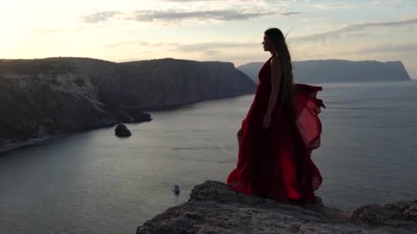 Uma jovem vestida com um vestido vermelho fica ao lado do mar em uma rocha, Uma menina desfruta da vista do mar e do nascer do sol, seu cabelo é soprado pelo vento, a bainha do vestido está balançando. — Vídeo de Stock