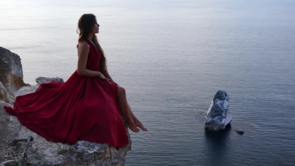 Młoda kobieta ubrana w czerwoną sukienkę siedzi nad morzem na skale, dziewczyna cieszy się widokiem na morze i wschód słońca, jej włosy wieją na wietrze, obszycie sukienki rozwija. — Wideo stockowe