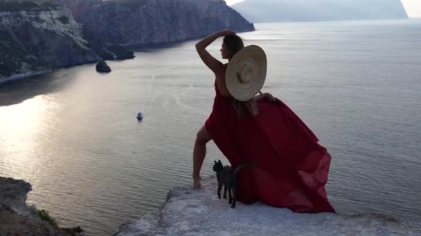 Uma jovem vestida com um vestido vermelho fica ao lado do mar em uma rocha, Uma menina desfruta da vista do mar e do nascer do sol, seu cabelo é soprado pelo vento, a bainha do vestido está balançando. — Vídeo de Stock