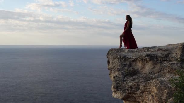 Uma jovem vestida com um vestido vermelho fica ao lado do mar em uma rocha, Uma menina desfruta da vista do mar e do nascer do sol, seu cabelo é soprado pelo vento, a bainha do vestido está balançando. — Vídeo de Stock