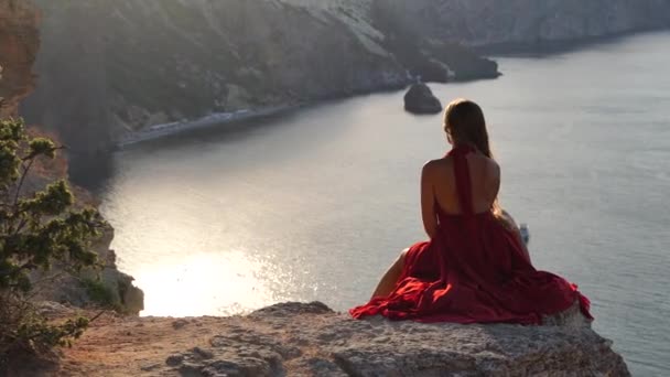 Uma jovem vestida com um vestido vermelho está sentada ao lado do mar em uma rocha, Uma menina está apreciando a vista do mar e do nascer do sol, seu cabelo está soprando no vento, a bainha do vestido está se desenvolvendo. — Vídeo de Stock