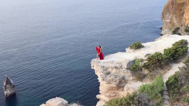 Uma jovem vestida com um vestido vermelho fica ao lado do mar em uma rocha, Uma menina desfruta da vista do mar e do nascer do sol, seu cabelo é soprado pelo vento, a bainha do vestido está balançando. — Vídeo de Stock