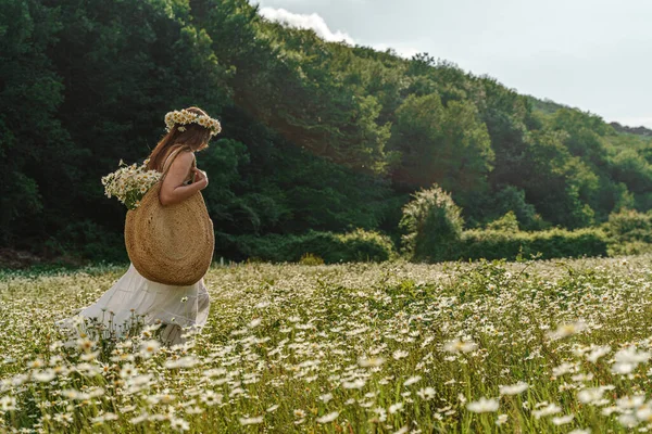 A young woman in a white sundress, a wreath of daisies with a large wicker bag on her shoulder is walking through a field of daisies, against the background of the forest, the sunset light