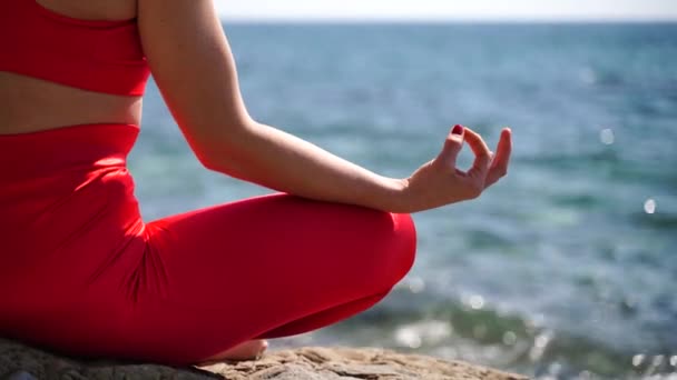 Uma jovem mulher de sportswear vermelho relaxa enquanto pratica ioga na praia pelo mar calmo, close-up de mãos, mudra gyan e pose de lótus. — Vídeo de Stock