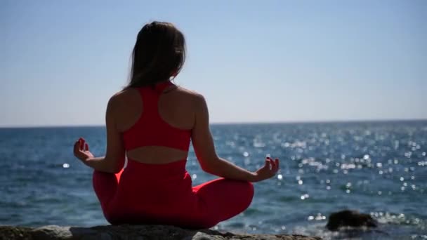 Una joven en ropa deportiva roja se relaja mientras practica yoga en la playa junto al mar tranquilo, primer plano de las manos, gyan mudra y pose de loto. — Vídeos de Stock