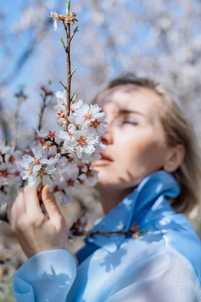 Retrato de una joven rubia en un floreciente jardín de primavera. Los almendros están floreciendo. La mujer está vestida con ropa azul.. —  Fotos de Stock