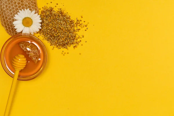 Ένα κίτρινο κουτάλι μέλι βουτηγμένο σε ένα βάζο με μέλι και ψωμί μέλισσας σκορπισμένο σε κίτρινο φόντο διακοσμημένο με ζωντανό χαμομήλι. Μέλι στάζει γύρω, ωραία και καλώντας φωτογραφίες. Έννοια υγιεινής διατροφής. — Φωτογραφία Αρχείου