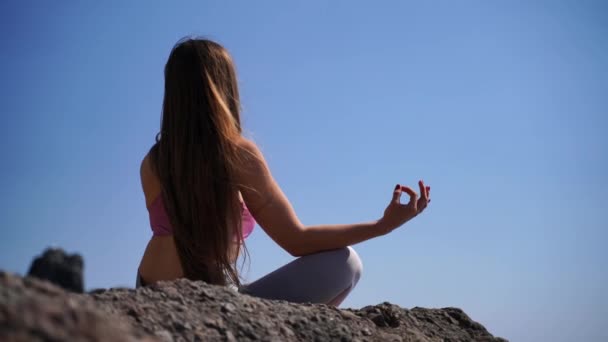 En ung kvinde i sportstøj slapper af, mens hun praktiserer yoga på stranden ved det rolige hav, tæt på hænder, gyan mudra og lotus udgør.. – Stock-video