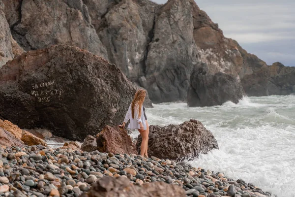 A beautiful girl in a white shirt and black swimsuit stands on the edge of a cliff, big waves with white foam. A cloudy stormy day at sea, with clouds and big waves hitting the rocks. — Stock Photo, Image