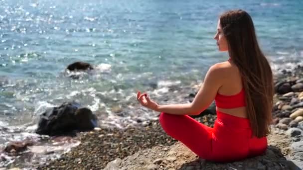 Une jeune femme en tenue de sport rouge se détend tout en pratiquant le yoga sur la plage au bord de la mer calme, gros plan des mains, mudra gyan et pose de lotus. — Video