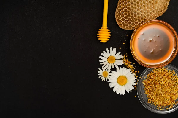 Un cucchiaio giallo di miele immerso in un barattolo di miele e pane d'api sparsi su uno sfondo nero decorato con camomilla viva. Miele gocciolante intorno, foto belle e invitanti. Concetto di cibo sano. — Foto Stock