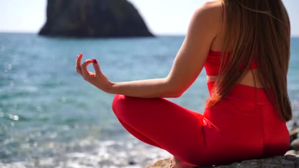 En ung kvinna i röda sportkläder slappnar av medan hon utövar yoga på stranden vid det lugna havet, närbild av händer, gyansk mudra och lotus utgör. — Stockvideo