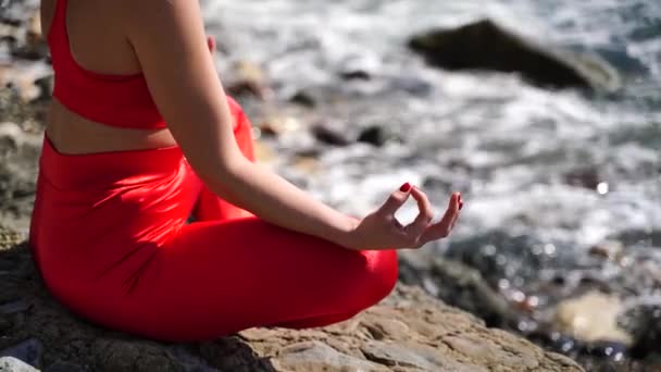 Une jeune femme en tenue de sport rouge se détend tout en pratiquant le yoga sur la plage au bord de la mer calme, gros plan des mains, mudra gyan et pose de lotus. — Video