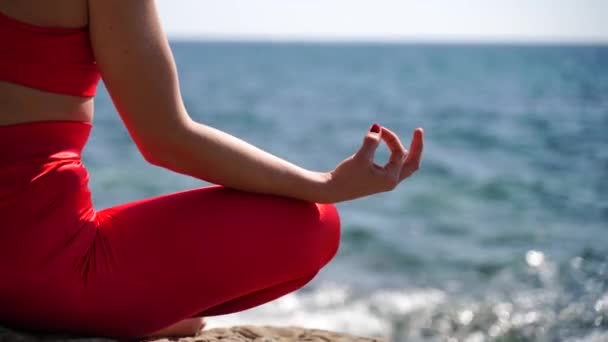 Une jeune femme en tenue de sport rouge se détend tout en pratiquant le yoga sur la plage au bord de la mer calme, gros plan des mains, mudra gyan et pose de lotus. — Video