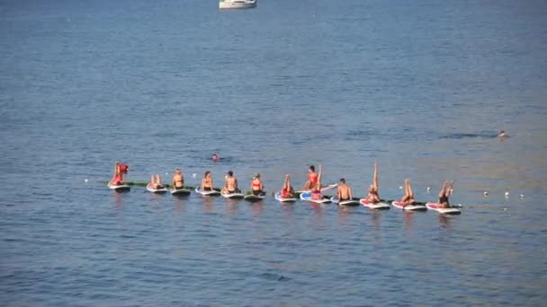 Un grupo de mujeres y hombres en trajes de baño hacen yoga y pilates en una tabla de sup en el mar tranquilo, temprano en la mañana. El concepto de un estilo de vida saludable. — Vídeos de Stock