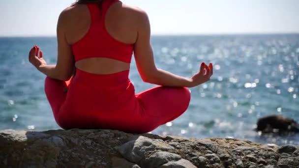 Una joven en ropa deportiva roja se relaja mientras practica yoga en la playa junto al mar tranquilo, primer plano de las manos, gyan mudra y pose de loto. — Vídeo de stock