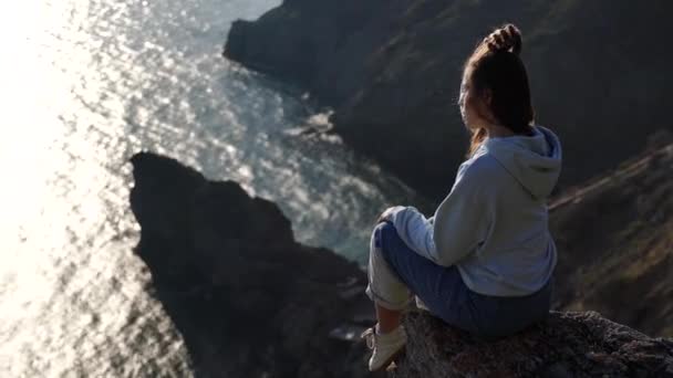 Vue arrière d'une jolie fille caucasienne en jeans et un sweat à capuche bleu se trouve sur une grande pierre au-dessus de la mer au coucher du soleil et regarde vers le bas vers le rivage. Dans le contexte de la côte de la mer, rochers dans le — Video
