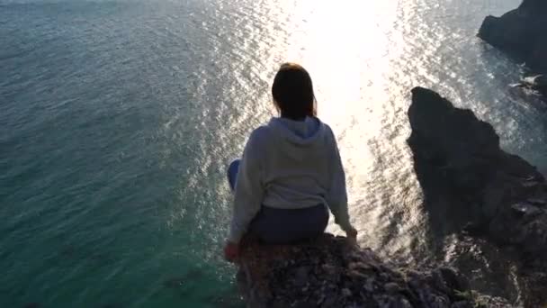 Vue arrière d'une jolie fille caucasienne en jeans et un sweat à capuche bleu se trouve sur une grande pierre au-dessus de la mer au coucher du soleil et regarde vers le bas vers le rivage. Dans le contexte de la côte de la mer, rochers dans le — Video