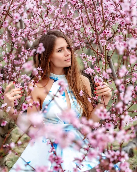 Çiçek açan şeftali ağacında oturan gülümseyen güzel kız beyaz-mavi elbiseli, uzun saçlı. Gülen surat. Bahar geldi.. — Stok fotoğraf