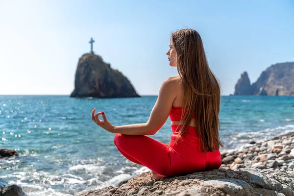 En ung kvinna i röda leggings och en röd topp med långt löst hår utövar yoga utomhus vid havet på en solig dag. Kvinnors yoga, kondition, pilates. Begreppet en hälsosam livsstil, harmoni. — Stockfoto