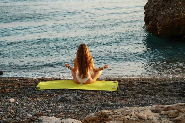 Beautiful woman in a bathing suit and long loose hair practicing yoga on the beach, Yoga Ardha Padmasana in the half-lotus position. — Stock Photo, Image