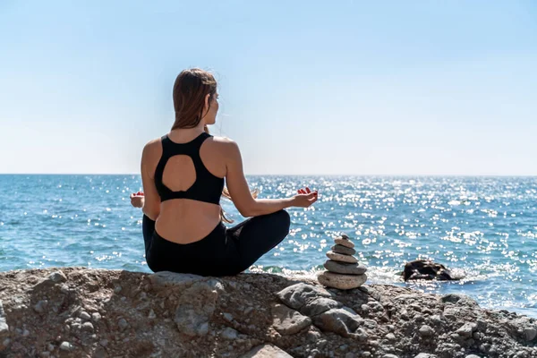 Junge Frau in schwarzen Leggings und schwarzem Top mit langen wallenden Haaren, die an einem sonnigen Tag im Freien am Meer Stretching übt. Frauen-Yoga, Fitness, Pilates. Das Konzept eines gesunden Lebensstils, Harmonie — Stockfoto