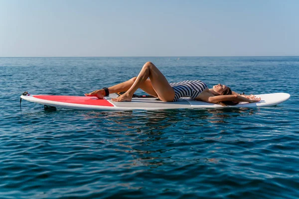 Fille sportive sur une planche de surf morve dans la mer par une journée d'été ensoleillée. En maillot de bain rayé, il s'allonge sur le dos. Activités estivales par Stortom by the sea — Photo