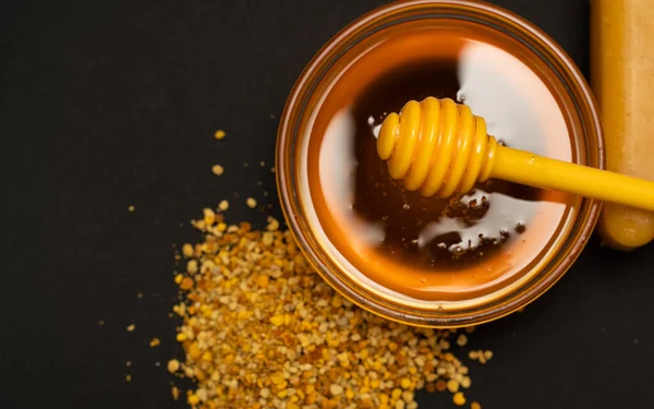 Ein gelber Löffel Honig in einem Glas Honig und Bienenbrot auf schwarzem Hintergrund verstreut. Honig tropft herum, schöne und einladende Fotos. Konzept für gesunde Ernährung. — Stockfoto