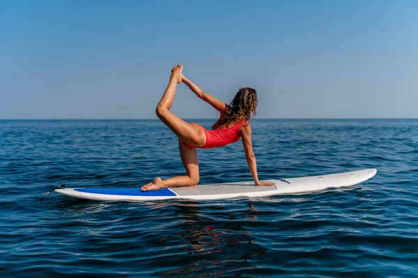 Sportliches Mädchen auf einem Surfbrett im Meer an einem sonnigen Sommertag. Im roten Badeanzug sitzt sie im Spagat. Sommerunterhaltung auf Stortom am Meer — Stockfoto