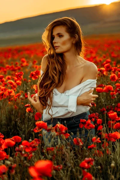 Bela jovem mulher de cabelos longos em um campo de papoula ao pôr do sol com cabelo voador. Vestido com uma camisa branca e saia jeans. — Fotografia de Stock