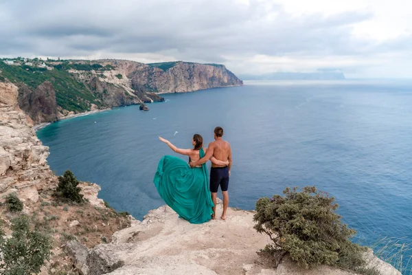 Une belle fille en robe verte et des hommes courent à la mer, sur fond de grands rochers et de la mer — Photo