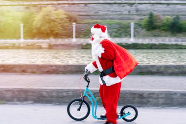 Noel Baba 'nın hediye dolu açık bir araba bagajıyla yan portresi hediyeleri dağıtmak için büyük tekerlekli bir çocuk scooter' ı sürüyor ve 2020 yılındaki kovid19 Coronavirus salgınından dolayı maske takıyor.