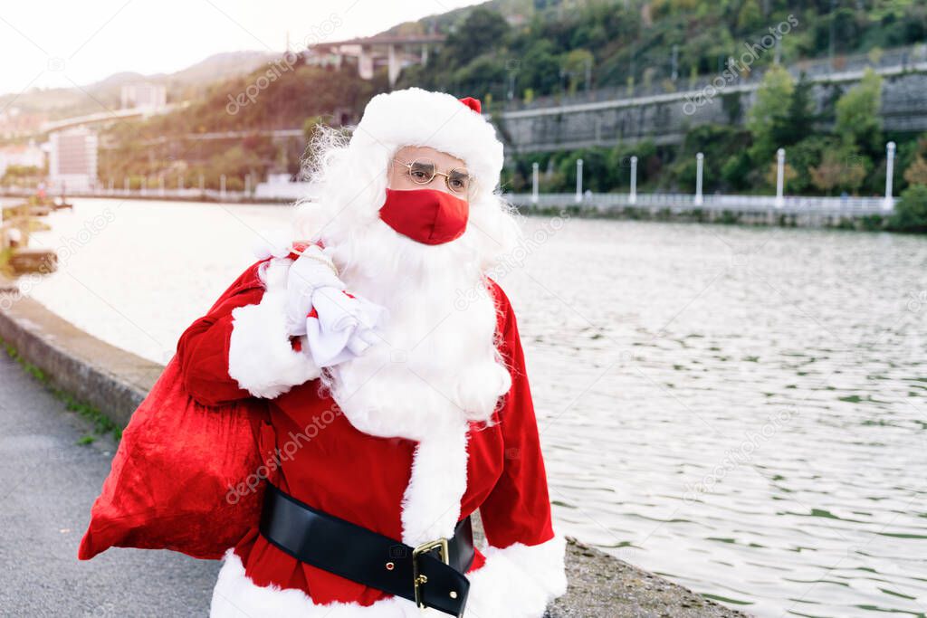 Portrait of Santa Claus walking down the street with a gift bag on his shoulder because he is carrying gifts to children with red face masks due to the covid19 coronavirus pandemic on Christmas 2020
