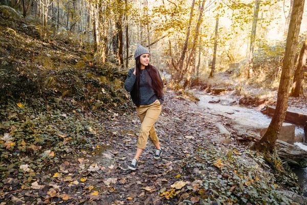 very beautiful and very happy woman in the autumn season walking through a forest with the leaves of the orange and yellow trees and a river. The woman is wearing a black coat and a gray wool cap