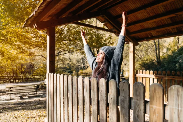 Portrait of a very beautiful and very happy woman in the autumn season with her arms raised in a forest hut with all the leaves of the orange and yellow trees. The woman is wearing warm clothes