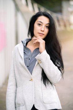 Çenesine dokunan, uzun siyah saçlı, kentli, yönetici ya da öğrenci tarzı kıyafetlerle sokakta poz veren güzel beyaz bir kadının moda portresi.