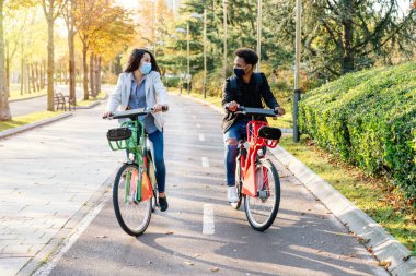2020 Covid19 Coronavirus salgını için maske takan iki genç adam ve kadının önünde gün batımında birçok ağacın olduğu güzel bir parkta bisiklet paylaşıyorlar.