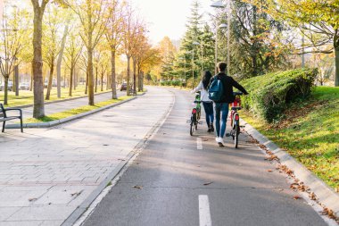 İki genç gelin ve damat gün batımında birçok ağacın olduğu güzel bir parkta bisiklet paylaşıyorlar.
