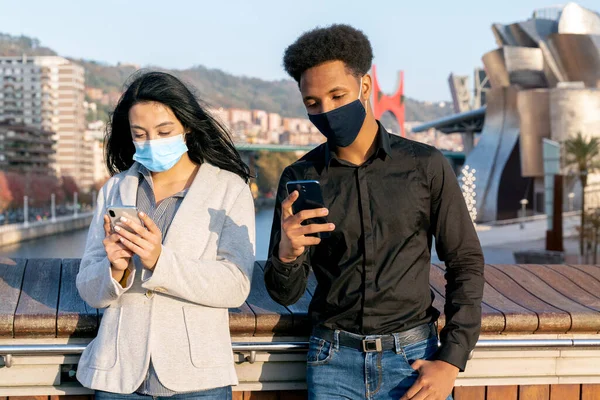 Portrait of a young couple on the street using their mobile phone writing text with a face mask due to the 2020 covid-19 coronavirus pandemic boy witn afro style hair