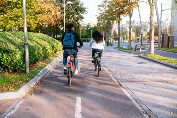 Arka kamerada iki genç adam ve bir kadın gün batımında birçok ağacın olduğu güzel bir parkta ortak bir elektrikli bisikletle bisiklet sürüyorlar.