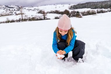 Sarışın kız karda oynuyor. Elleriyle kartopu yapıyor. Mavi kazak ve pembe şapka giyiyor.